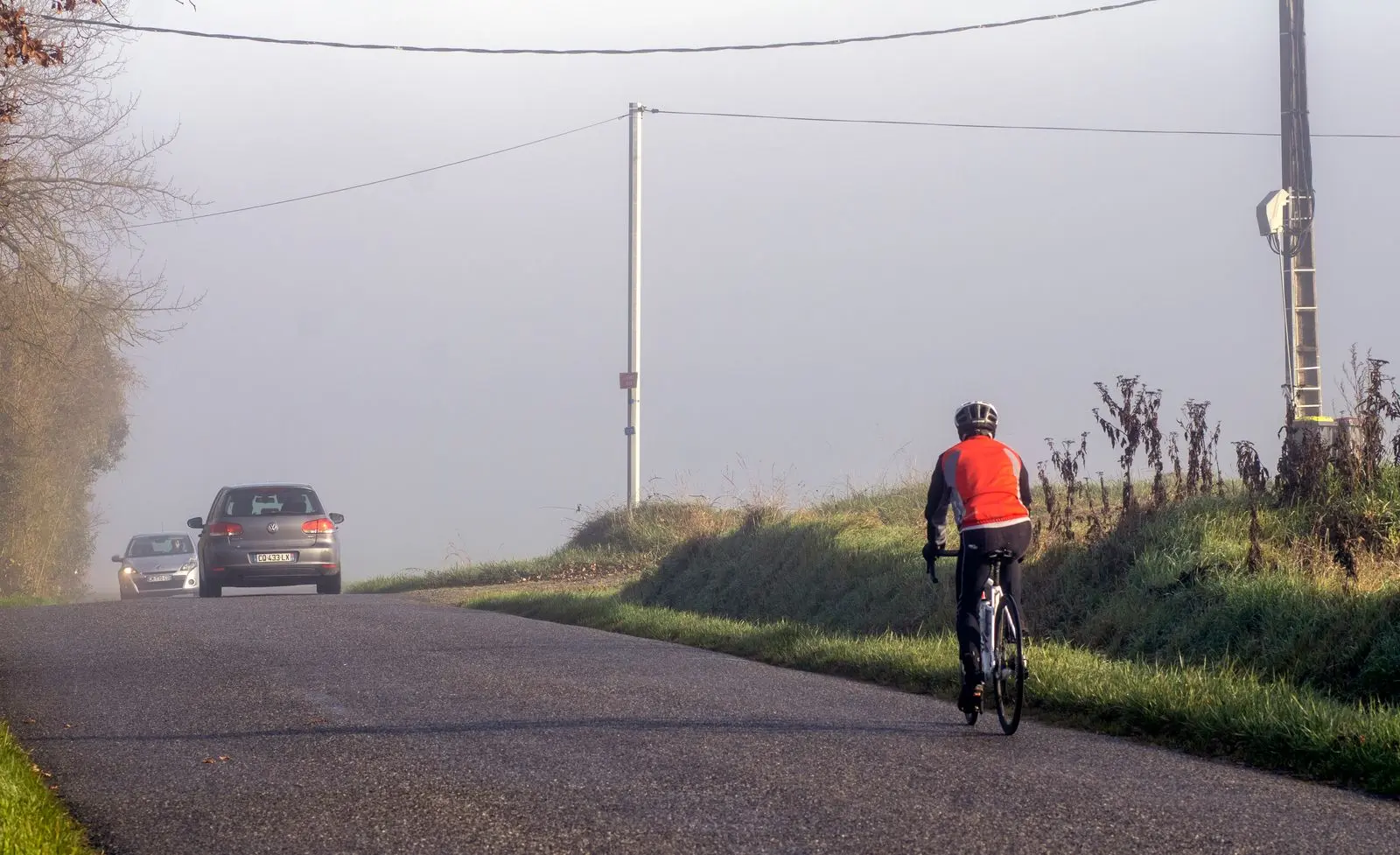 Ciclista con casco circulando por una carretera secundaria mientras se acercan dos coches